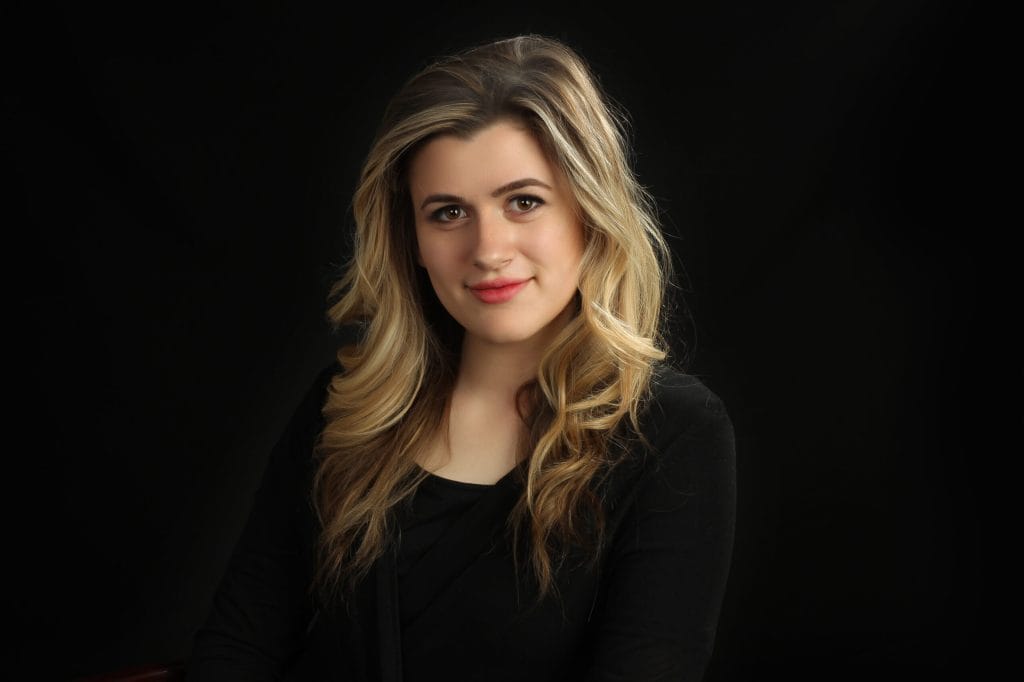 Smiling woman with long wavy blonde hair in black top, captured in a professional studio portrait with dark background