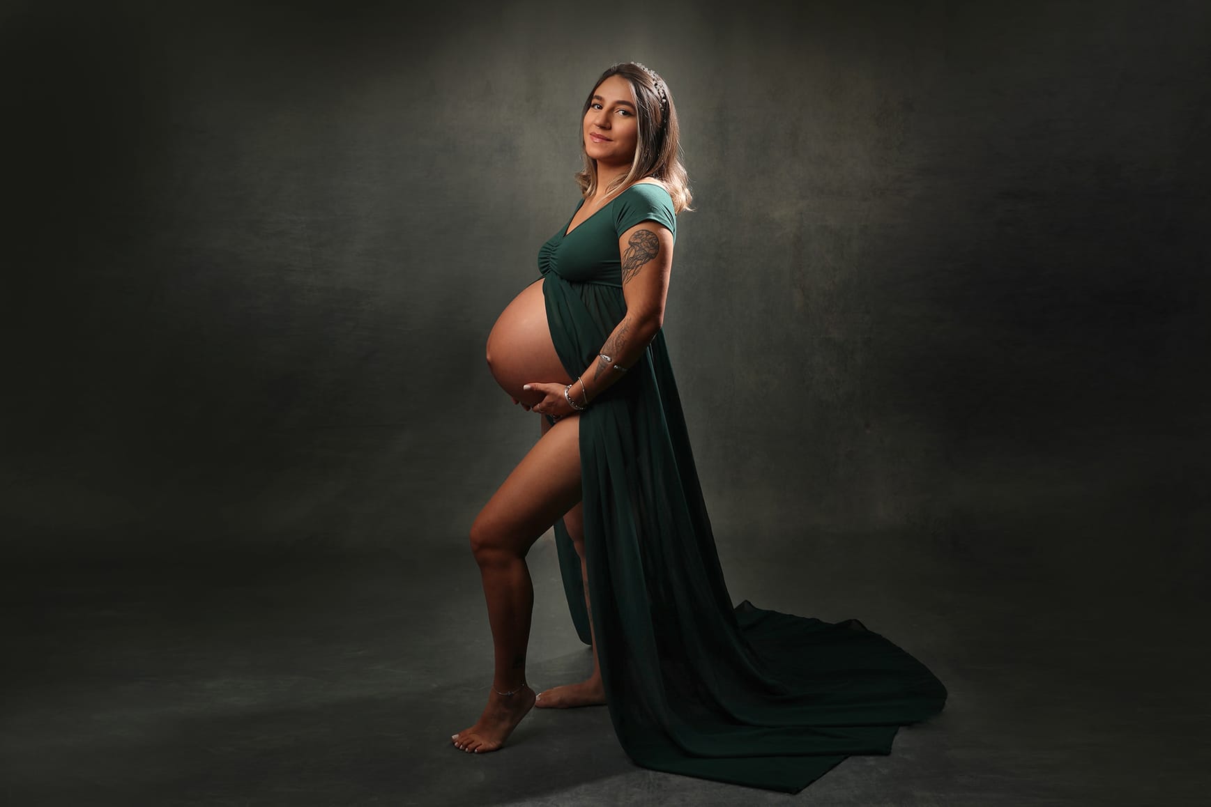 Pregnant woman in a long green gown cradles her bump against a dark background during a studio maternity photoshoot.