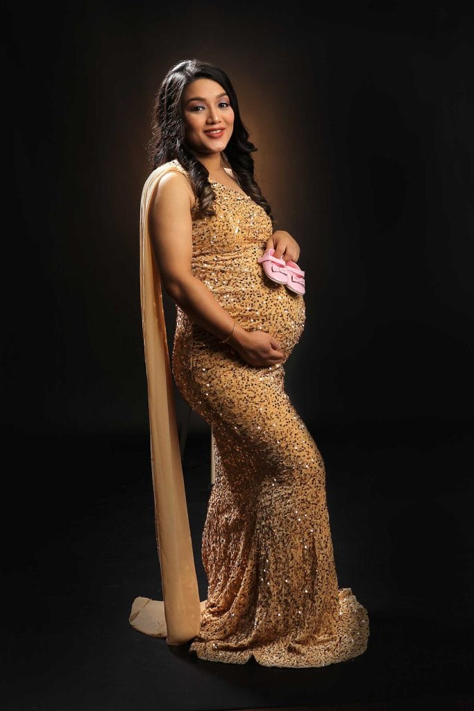 Pregnant woman in gold sequin gown holds pink baby shoes on her bump against a dark background in a maternity photoshoot.