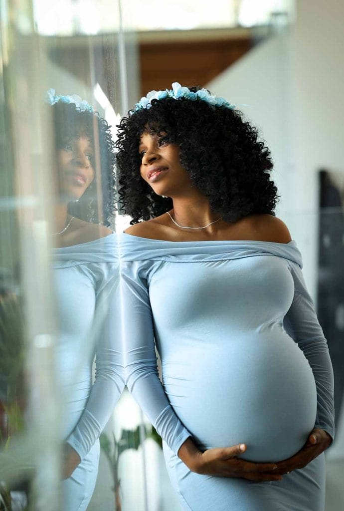 Pregnant woman in a blue dress and flower crown leans against a glass wall with reflection during a maternity photoshoot.