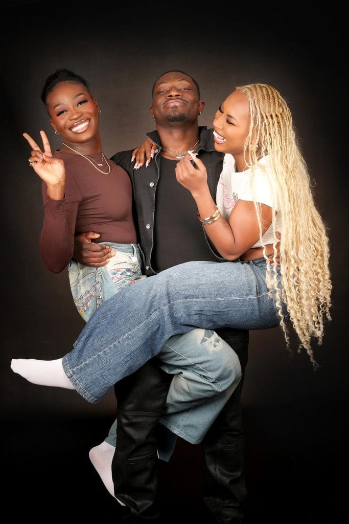 Man holding two smiling women in jeans; one woman gives a peace sign against a high-contrast black background.