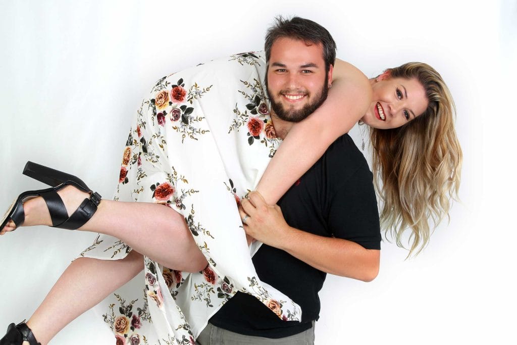 Smiling man carries woman in floral dress over his shoulder during a playful couple photoshoot on white background.