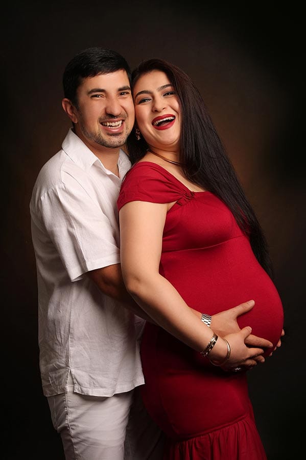 Laughing husband hugs pregnant wife in red dress from behind during a studio maternity photoshoot.