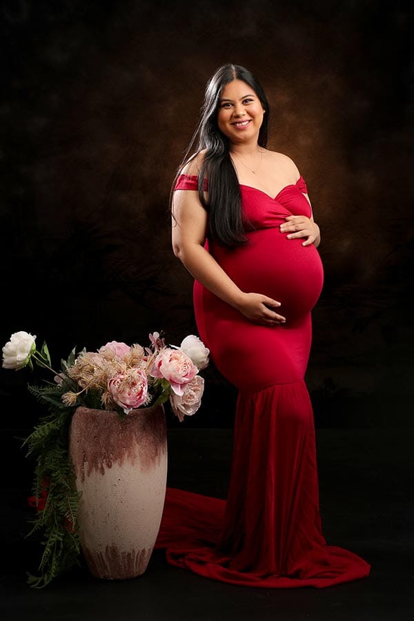 Pregnant woman in a red mermaid gown poses beside a floral arrangement for a studio Maternity Photoshoot.