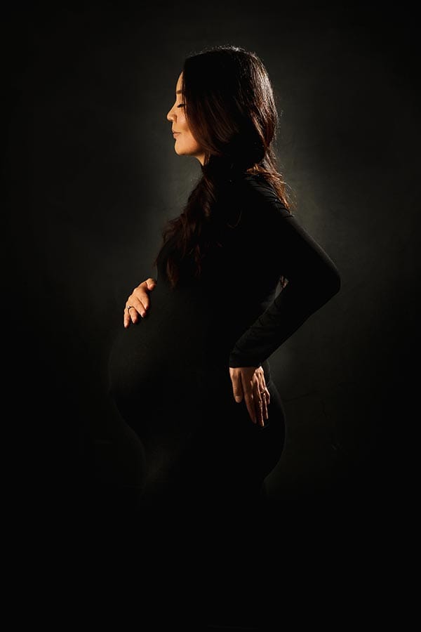 Pregnant woman in black dress stands in profile holding her bump against a dark background during a maternity photoshoot.