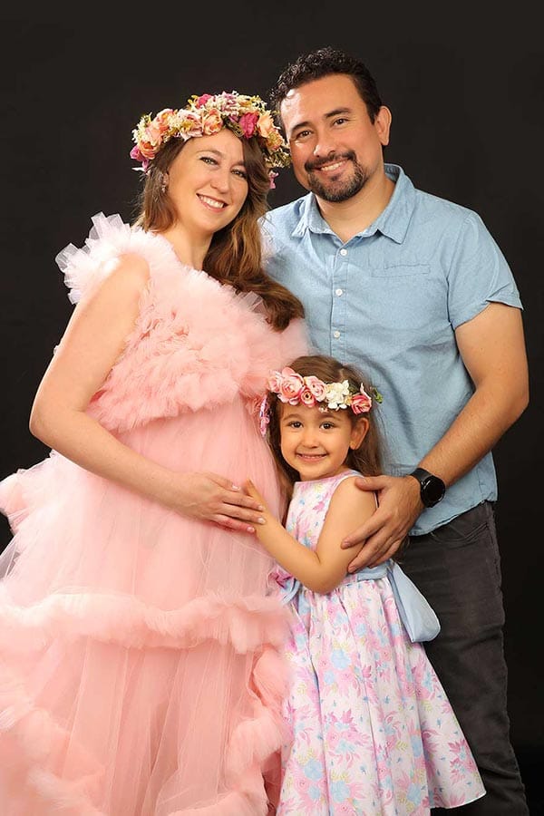Pregnant woman in pink dress smiles with husband and daughter wearing flower crowns during a family maternity photoshoot.