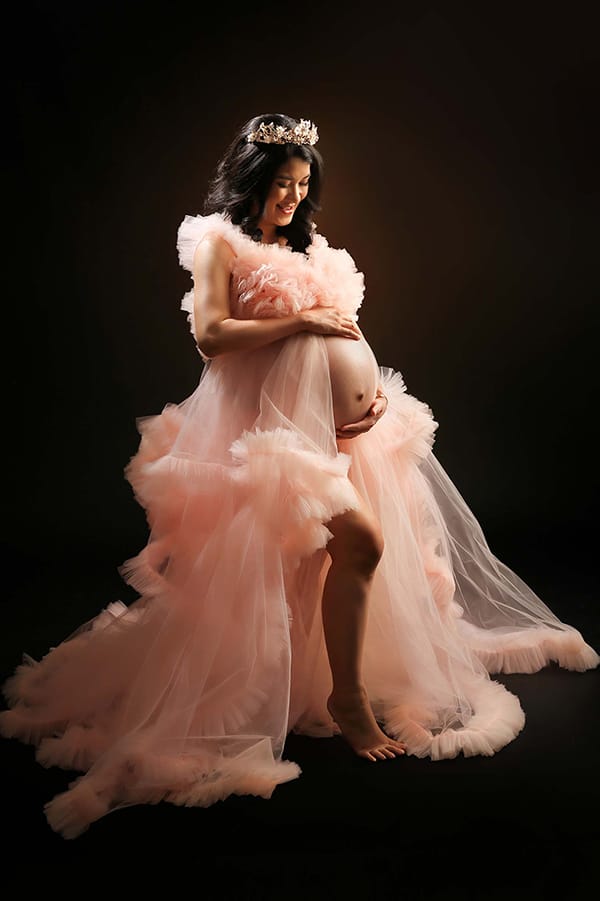 Pregnant woman in a pink ruffled gown and crown holds her bump against a dark background during a maternity photoshoot.