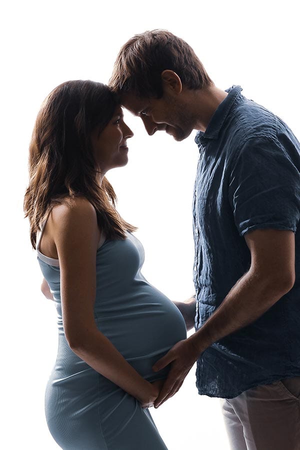 Pregnant woman in blue dress and husband touch foreheads against a white background during a studio maternity photoshoot.
