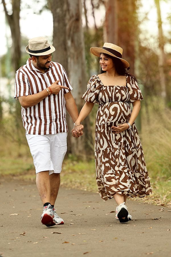 Smiling couple in hats walks holding hands while pregnant woman holds her bump during an outdoor maternity photoshoot.