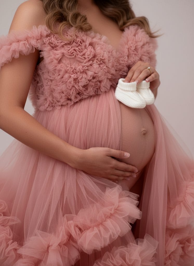 Pregnant woman in a pink ruffled gown holds small white baby shoes against her belly during a studio maternity photoshoot.